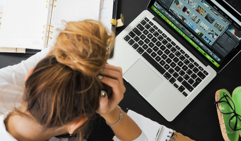 photo looking down on a woman at desk with a bun holding her head in her hands. A laptop and pair of glasses is sitting on the table in front of her.