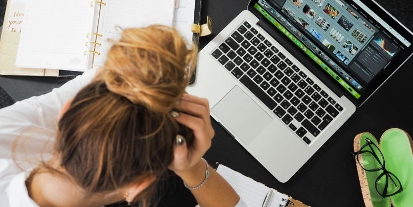 photo looking down on a woman at desk with a bun holding her head in her hands. A laptop and pair of glasses is sitting on the table in front of her.