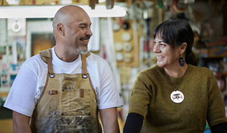 Photo of a man and a woman looking at each other and smiling, with a workshop in the background