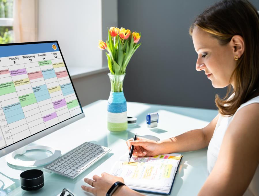 A young woman sitting at her computer with a notepad in front of her and a vase of flowers in the background