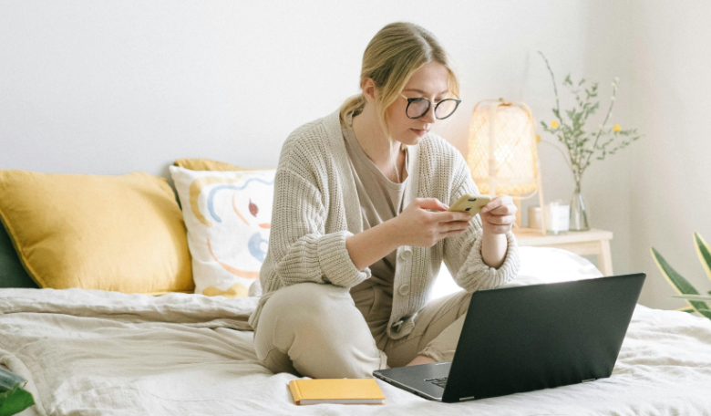 Woman sitting on her bed with a laptop and phone